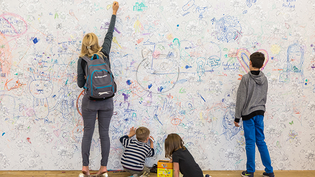 A lady and three children draw on a white wall, which already has a lot of drawings on it.
