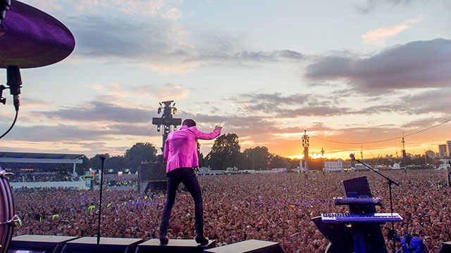 A man in a bright pink jacket performing on stage in Hyde park in front of thousand of people at sunset