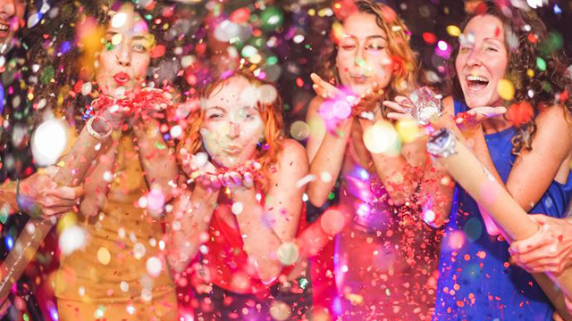 Four women, dressed in colourful dresses, celebrate as ticker tape falls around them.