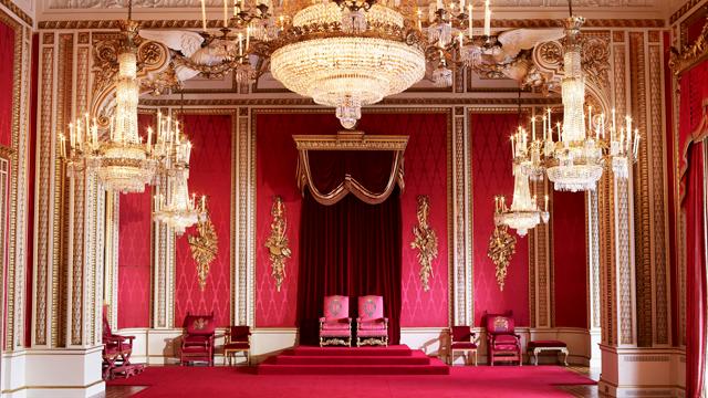 The Throne Room at Buckingham Palace, featuring two red throne chairs, red carpet and chandeliers.