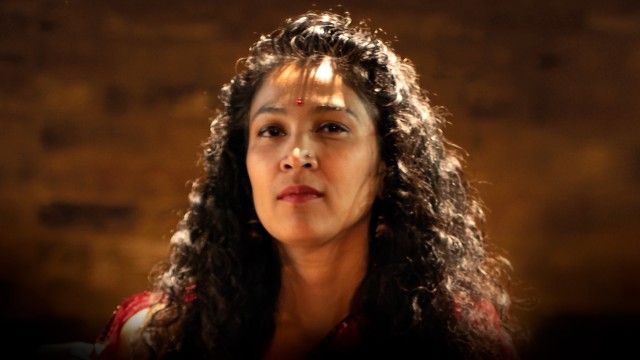 South Asian woman with curly hair wearing red lipstick and red bindi against a blurry brick background.
