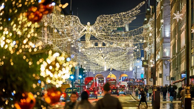 Un arbre de Noël avec des lumières blanches éblouissantes et des ornements rouges au premier plan. Des bus rouges circulent  sous des "anges" de Noël et autres décorations.