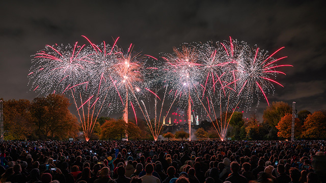 Red and white fireworks explode above a large crowd of people at night, with Battersea Power Station in the background.