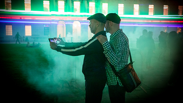 Two people take a selfie during Borealis, with strips of green, red and purple light and an illuminated mist in the background.