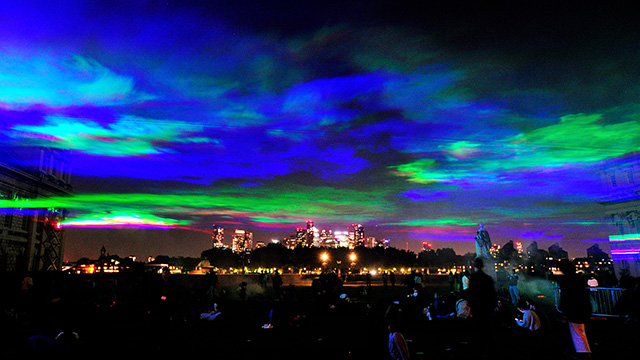 Blue, green and pink lights illuminate the skies at night above London's skyline, with skyscrapers lit up in the background, and a crowd of people in the foreground.
