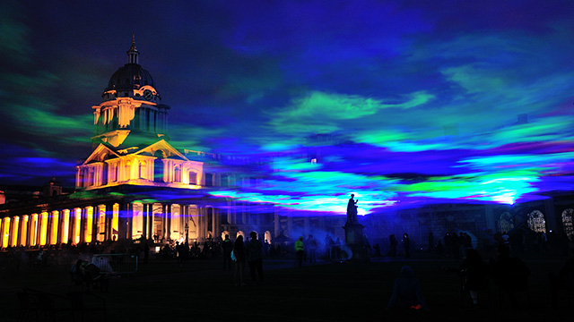 Blue and green lights illuminating the clouds above Greenwich at night, with a crowd of people and the silhouette of a statue below.