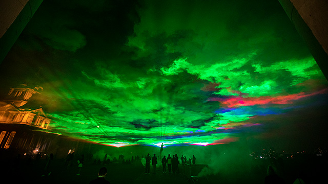 Green, red and blue lights illuminate the sky at night, with a crowd of people below.