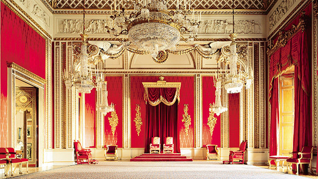 The Throne Room at Buckingham Palace with two thrones, red wallpaper and ornate gold decorations, including a chandelier.