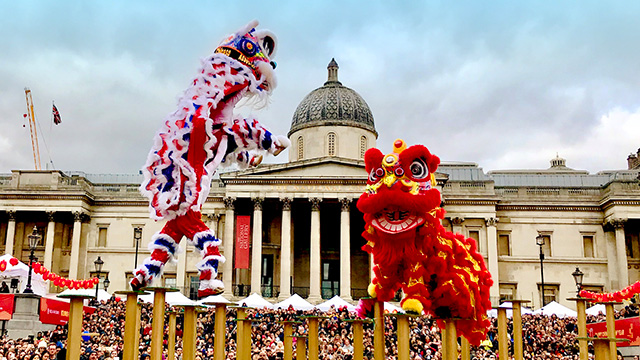 Two lion dance teams balance on yellow poles in Trafalgar Square, with crowds of people and the National Gallery behind.