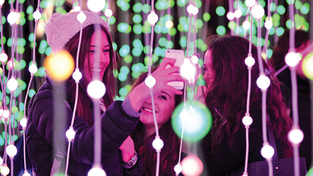 Three women take a selfie among strings of green and white fairy lights at night.