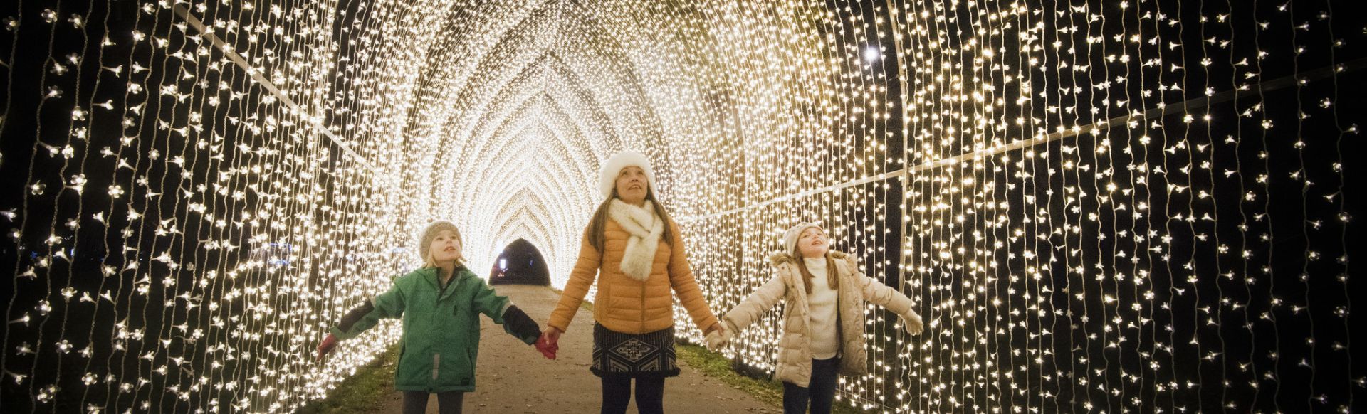 A family wearing coats and scarves hold hands as they stand under a bright yellow cathedral of lights at Christmas at Kew.