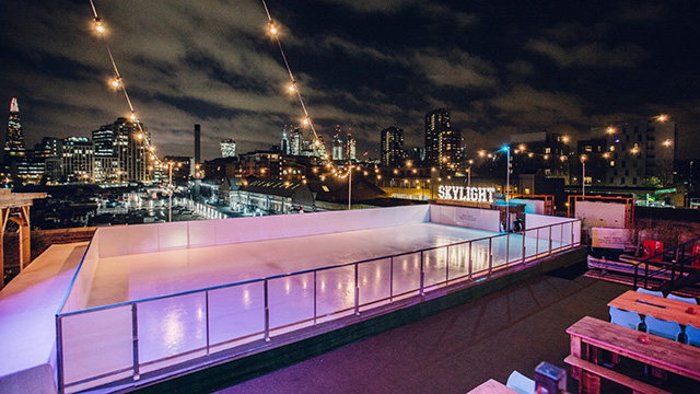 La patinoire du toit Skylight le soir, avec des lumières féeriques au-dessus de la patinoire et une vue sur Londres. 
