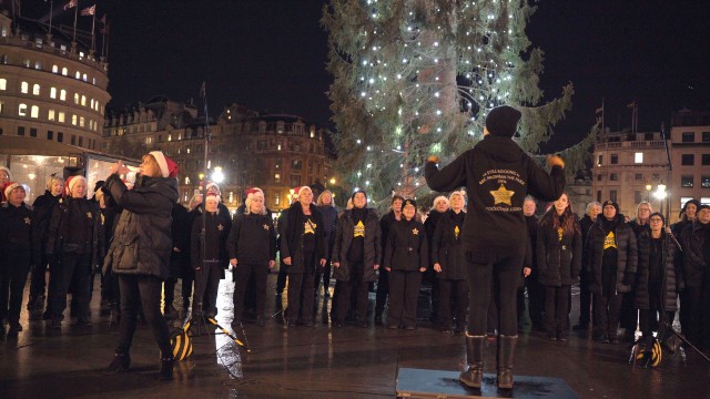 Carol singers in matching black clothes and Santa hats, led by a director, standing and singing in front of a festively decorated Trafalgar Square Christmas tree, covered in twinkling, vertical lights.