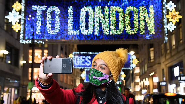 A lady wearing a face covering and a yellow bobble hat takes a selfie of the blue and yellow Oxford Street lights, which read "TO LONDON".