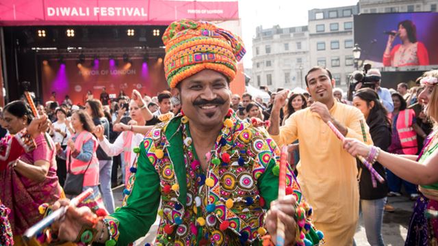 A man dressed in traditional clothing with flower garlands around his neck, at the Diwali festival, while other performers dance in the background.