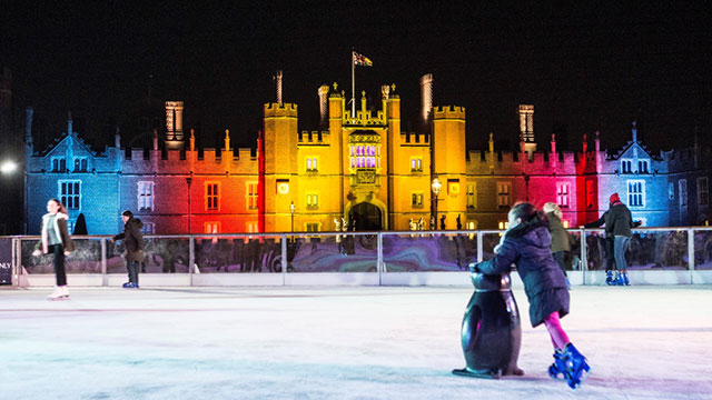 A young girl skating with a penguin on the ice rink at night time in front of Hampton Court Palace lit up in red, yellow and blue lights