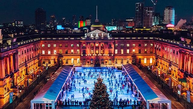 A aerial view of Somerset House ice rink at night with people ice skating and the city skyline behind
