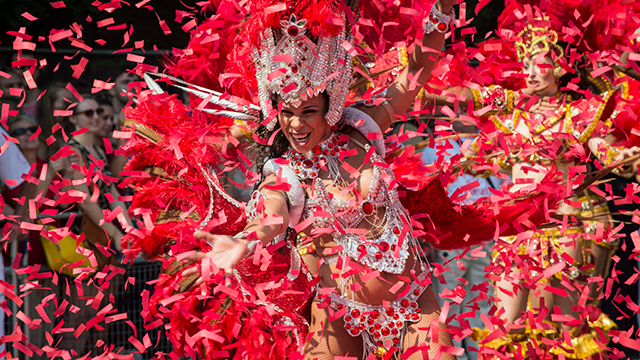 A woman dressed in a carnival headdress and clothing dances as red ticker tape fills the air at Notting Hill Carnival in London.