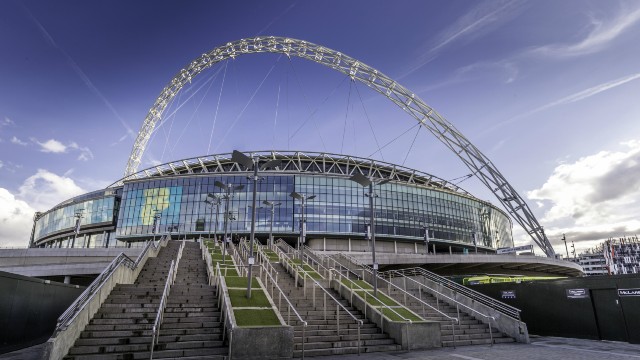 The outside of Wembley Stadium viewed from the bottom of the entrance steps with the venue's iconic arch emphasised against the blue sky.