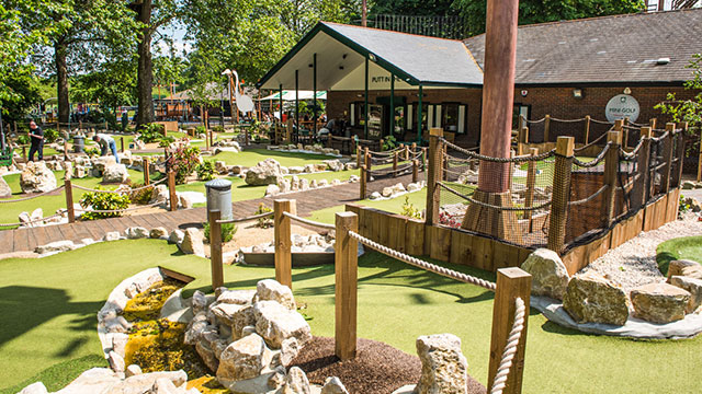 A mini-golf course set in a park with a clubhouse behind on a sunny day.