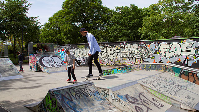 Two boys on skateboards gliding over the graffitied concrete ramps at Alexandra Palace skatepark on a sunny day.