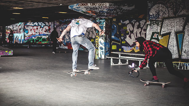 Two skateboarders performing tricks in the iconic concrete and graffitied Southbank Skate Space in London.