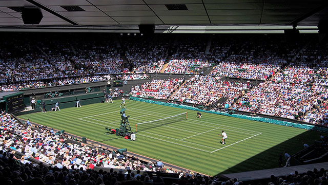 A tennis match being played on grass in front of an audience during summer in London