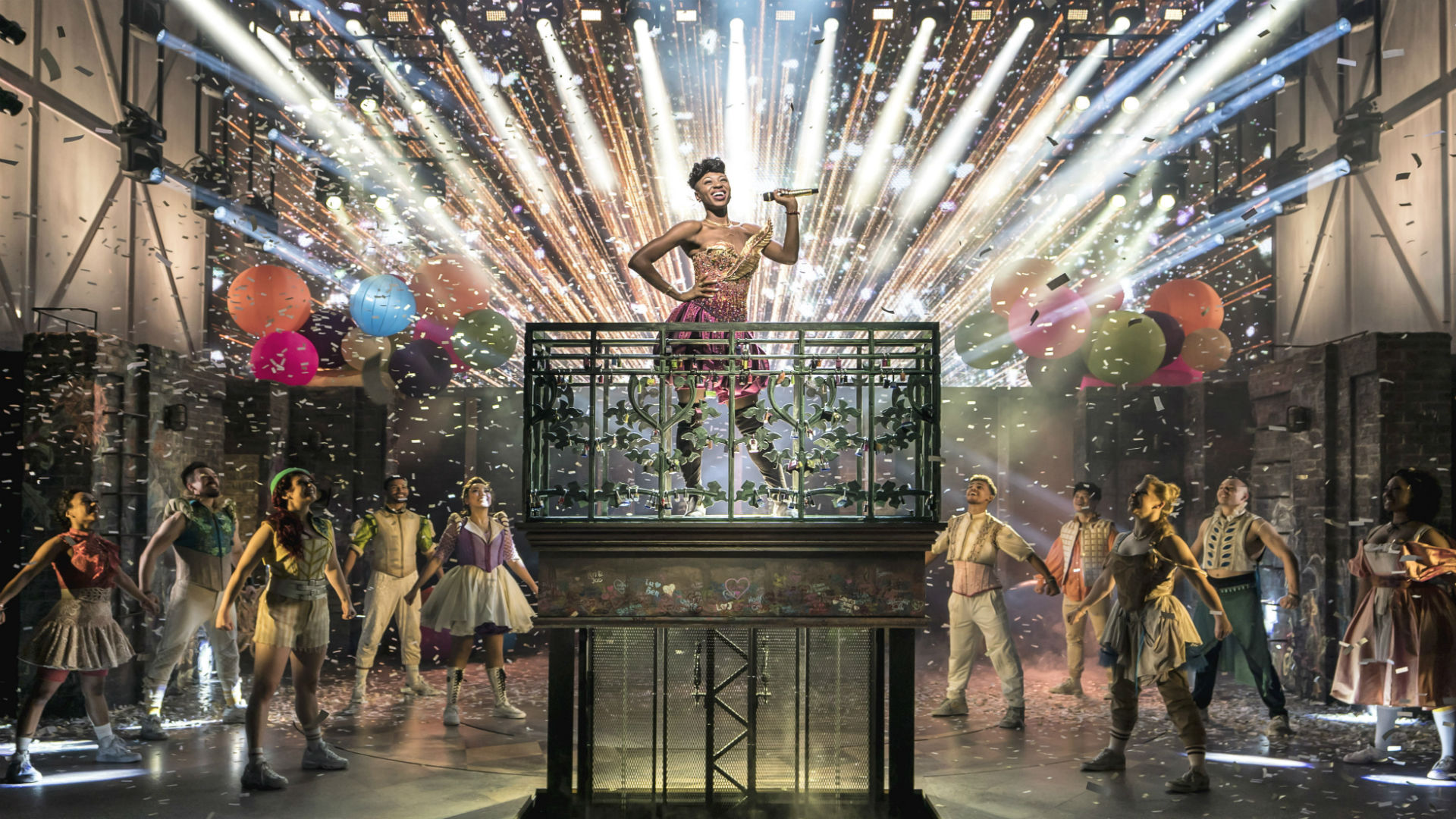 Juliet stands on a raised platform in the middle of the stage surrounded by cast members, coloured balloons, lights and ticker tape. 