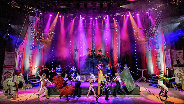 Des acrobates et artistes de cirque paradent sur scène sur un décor éclairé de néons de couleur violette.