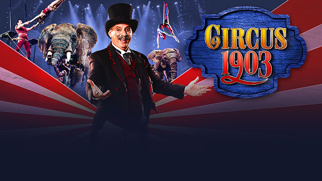 A man dressed in a black suit and hat with circus performers and an two elephant models in the background.