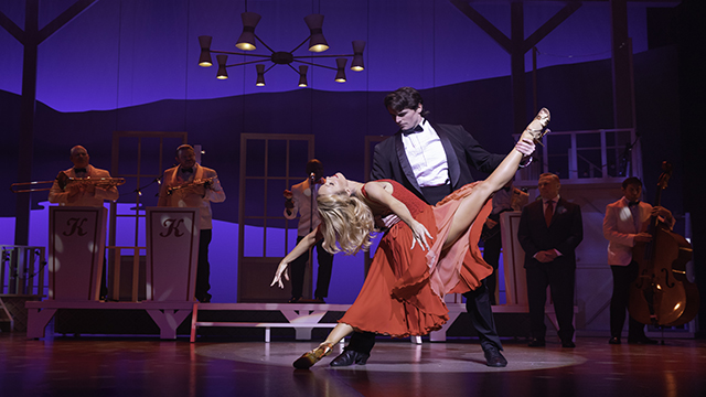 Un couple exécute un numéro de danse sur scène, avec des danseurs tous deux habillés pour l'occasion et un orchestre jouant en arrière-plan.