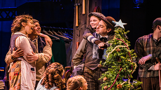 A group of three adults and three children are looking at each other as they sing around a green and sparkling christmas tree at the dolly parton christmas carol musical.