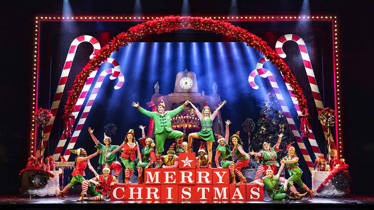 The cast of Elf The Musical is on stage, all dressed as elves on a stage decorated with giant candy canes.