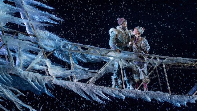 The actors playing Kristoff and Anna are standing on an ice bridge smiling at each other. 
