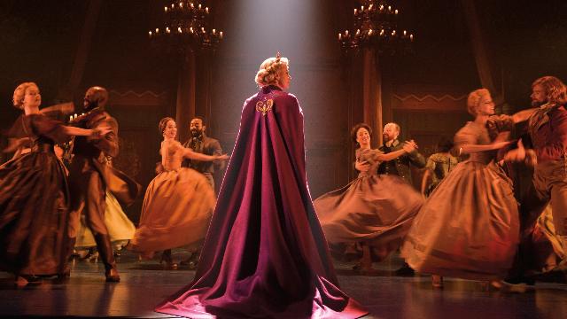 The actress playing Elsa on the musical Frozen is wearing a red cape and seems to have arrived at a ball where couples wearing gowns and three-piece suits are dancing around her.