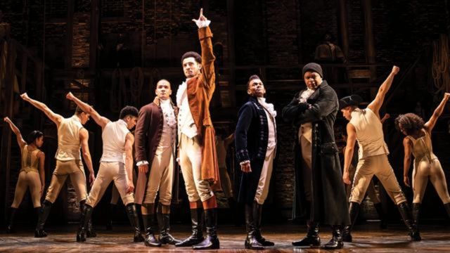A chorus of men dressed in brown and beige period outfits stand in a line with their arms in the air at Hamilton the musical.