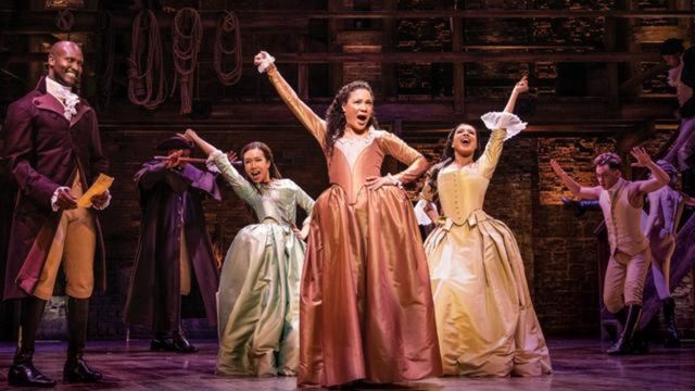 Three women, The Schuyler Sisters, stand together on stage with their arms in the air in large pastel dresses. n