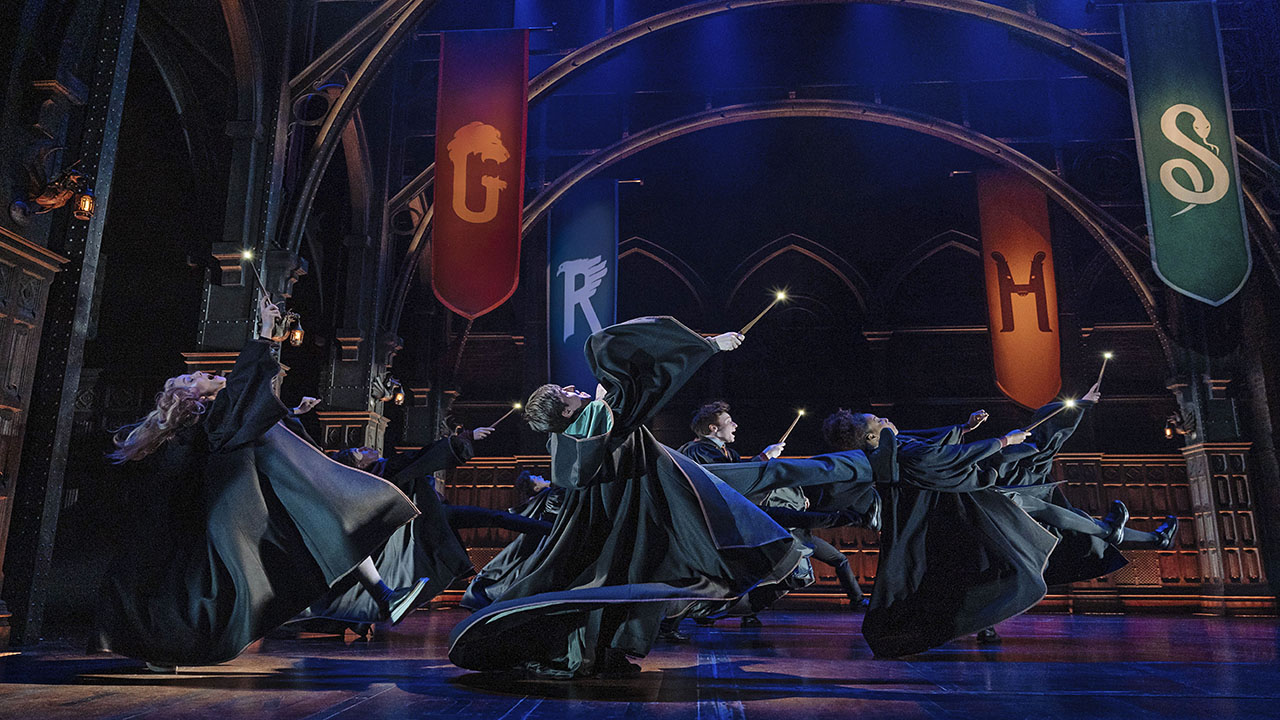 Hogwarts-Schüler in langen dunklen Roben tanzen auf der Bühne des Palace Theatre, über ihnen hängen die Fahnen der Häuser von Hogwarts.