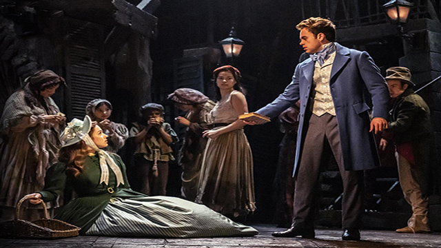 Die Darsteller tragen Renaissance-Kleidung, eine Frau mit grünem Mantel liegt auf dem Boden, ein Mann im blauen Mantel hält ein Buch in der Hand und im Hintergrund sind Menschen zu sehen.