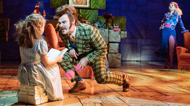 A girl dressed in white sits on some books, as a man dressed in green crouches near her, as part of the production of Matilda The Musical.