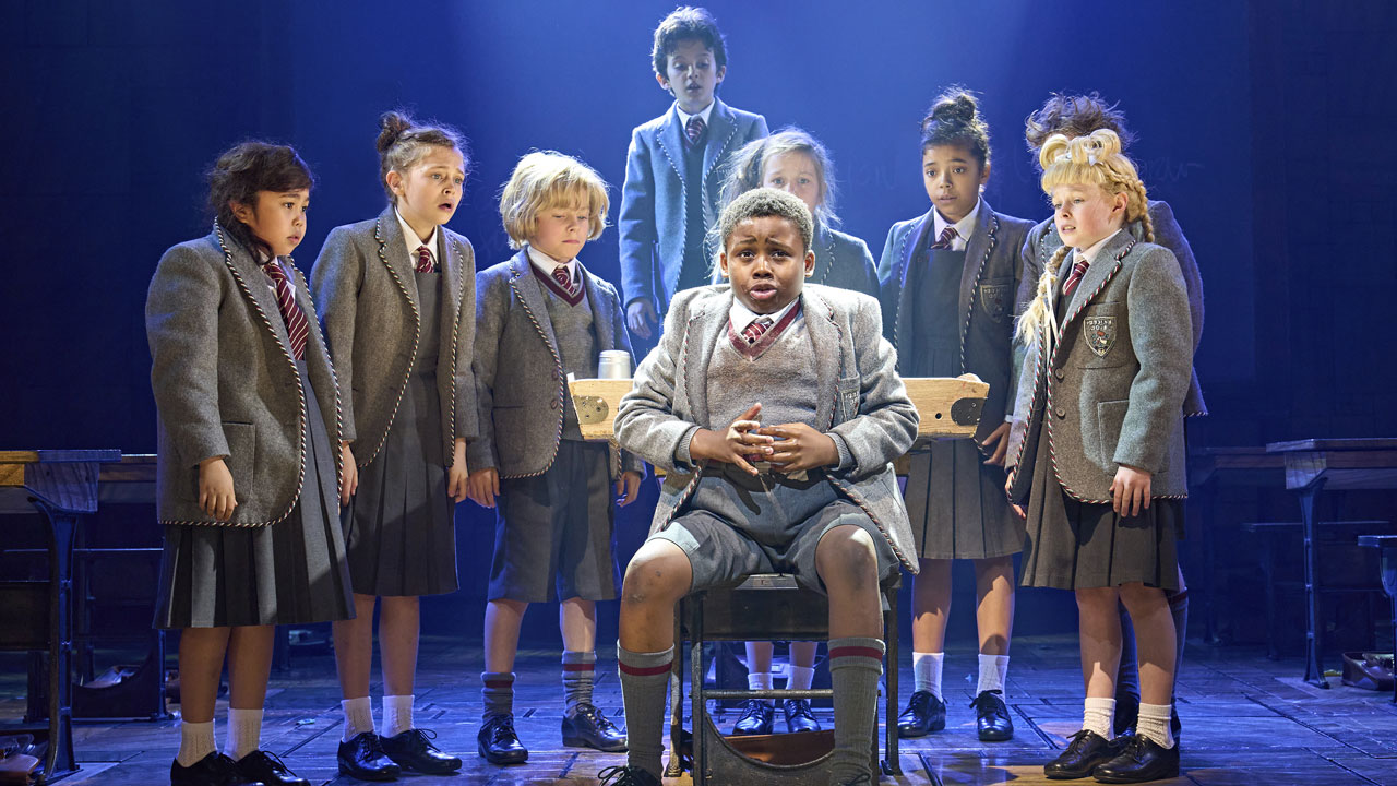 A group of school kids are surrounding one of their classmates seating on a chair during the show Matilda The Musical.