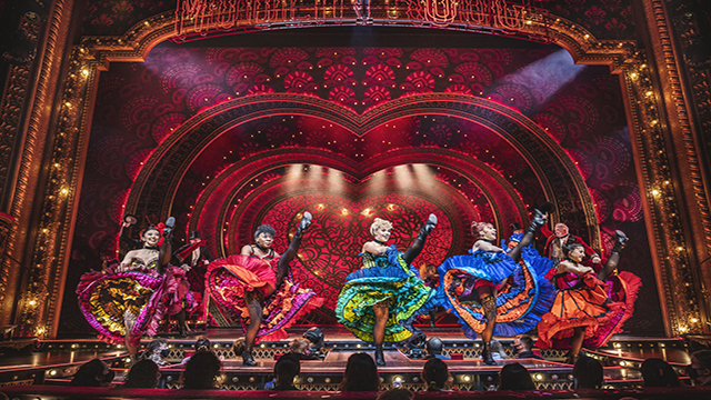 A troup of dancers wearing colourful dresses are dancing the French Cancan on the stage of Moulin Rouge The Musical in London.