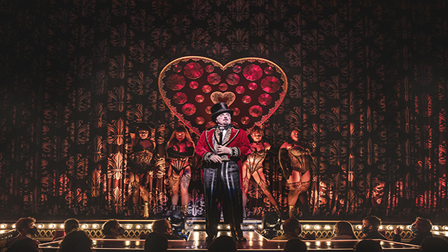 Harold Zidler, le propriétaire du Moulin Rouge, se tient sur le devant de la scène du Picadilly Theatre avec, derrière lui, quatre danseuses de cabaret vêtues de corsets en dentelle.