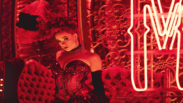 One of the dancers from the cast of Moulin Rouge The Musical is seating in a velvety boudoir under red neon lights and is wearing a lace corset.