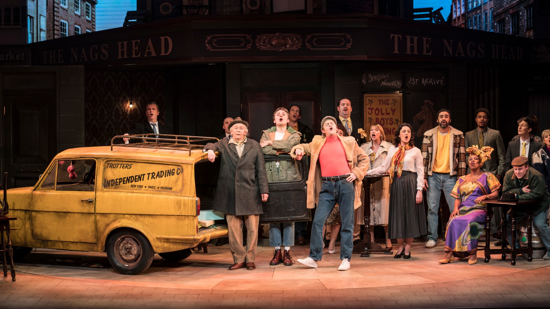 Die Darsteller von Only Fools and Horses singen auf der Bühne, mit einem gelben Auto auf der linken Seite des Bildes