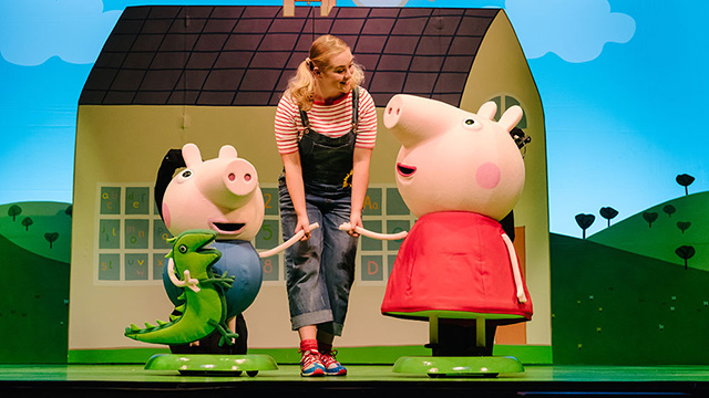A lady holds the hands of costumed characters Peppa Pig and George, who is carrying a dinosaur stuffed toy, in front of the school.
