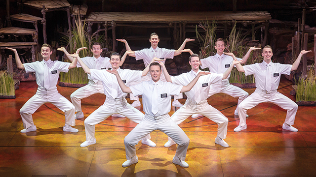 A group of male performers in white cruise outfits in pose on stage.