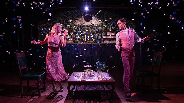 Ein männlicher und ein weiblicher Schauspieler, beide im Stil der 1920er Jahre gekleidet, tanzen in einem schwach beleuchteten Raum, in dem ein silbernes und blaues Rollband herabfällt und hinter dem eine Bar steht.