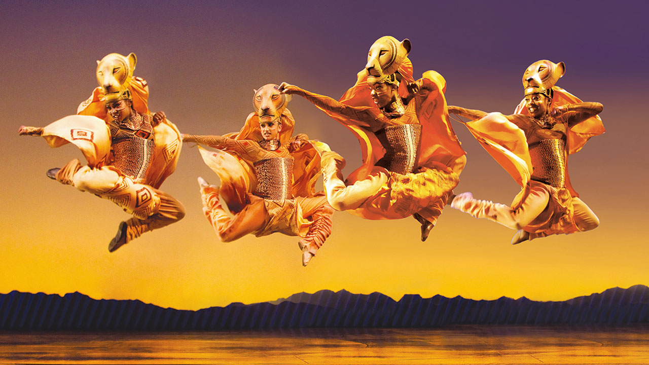 Four dancers leap across the stage in lion costumes in The Lion King on stage in the West End of London.