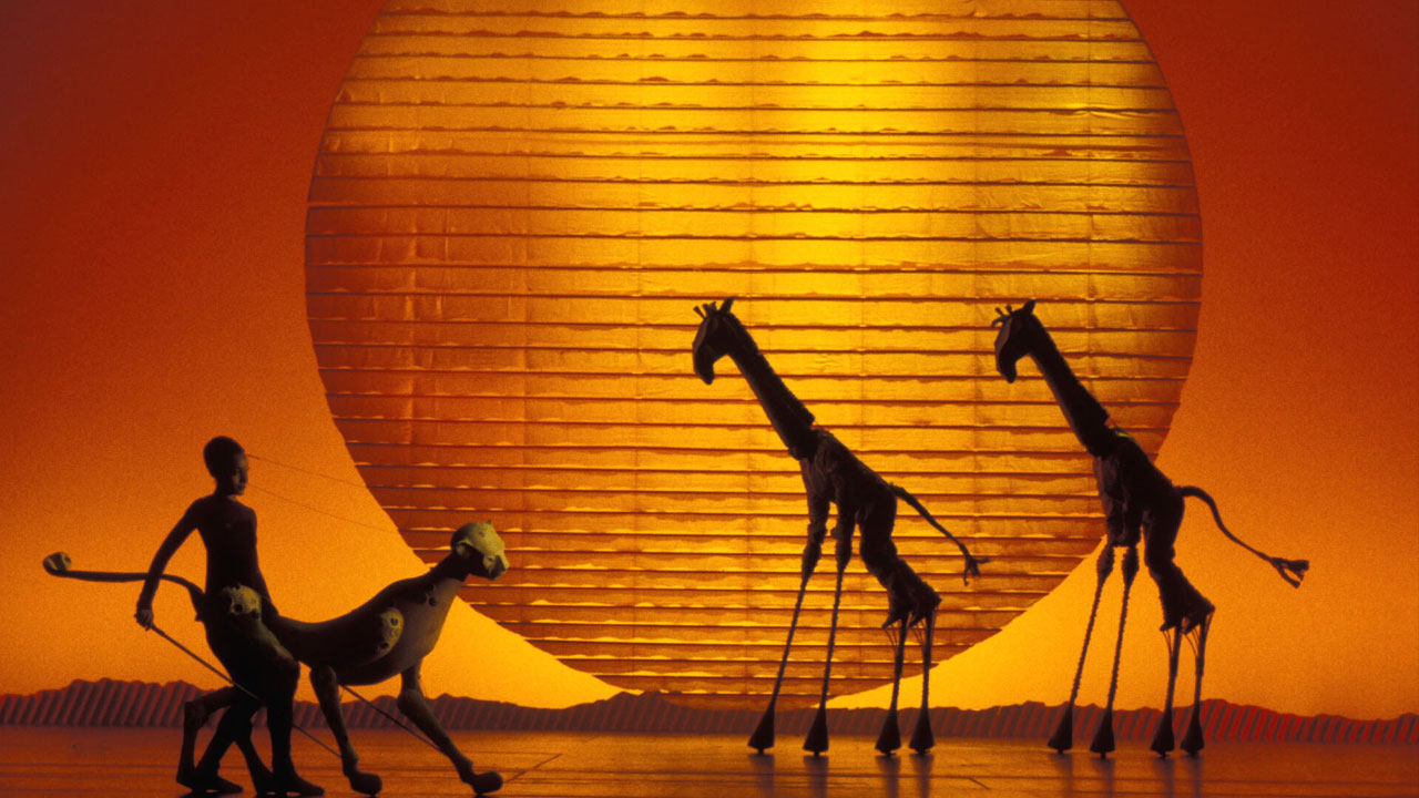Giraffe and Cheeter puppets on stage in front of a large yellow and orange sun in The Lion King